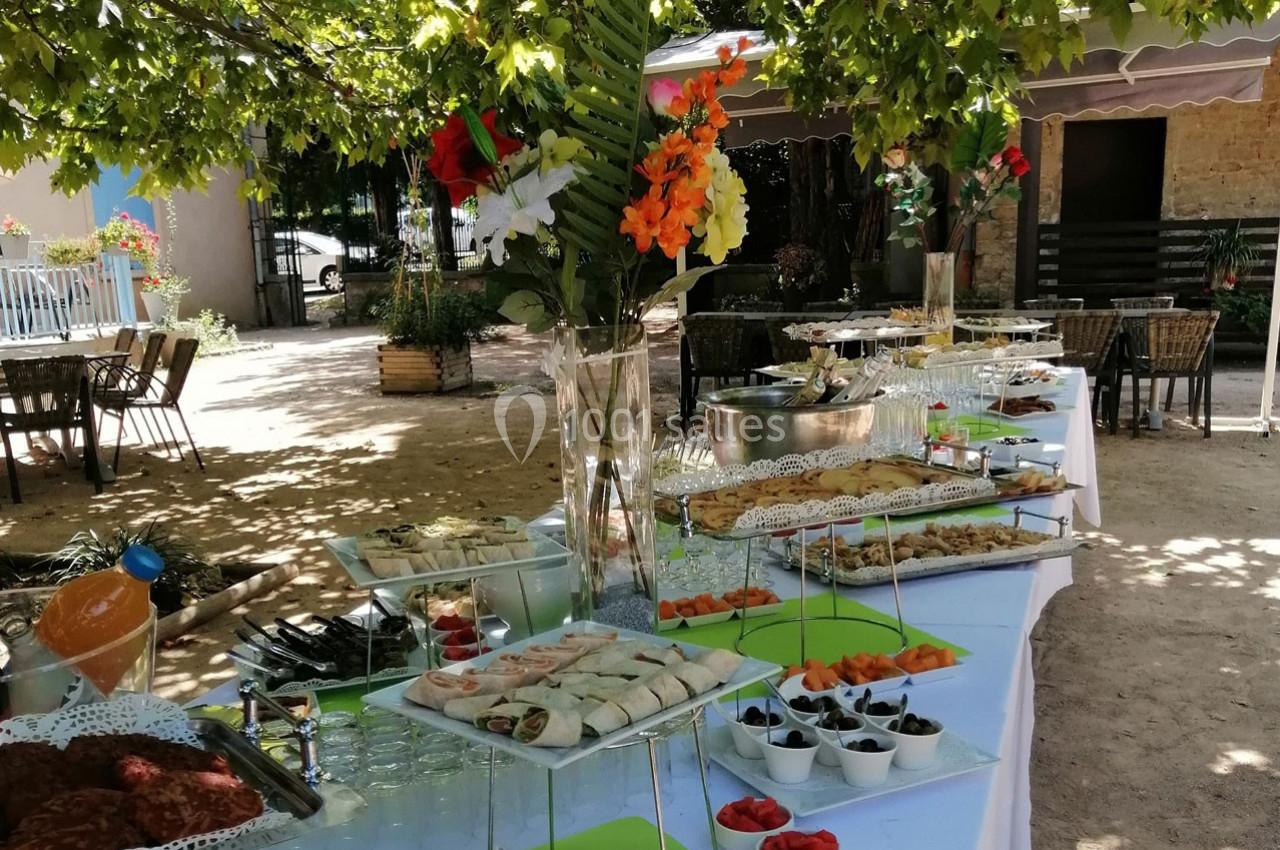 Buffet en extérieur sous des arbres, avec des plats variés, boissons et décorations florales sur des tables dressées.