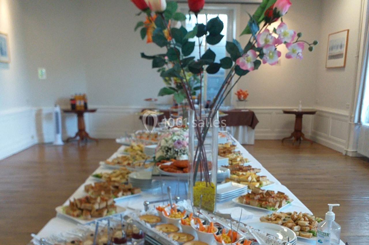 Table garnie d'un buffet varié avec plats salés et sucrés, décorée de fleurs au centre d'une salle lumineuse.