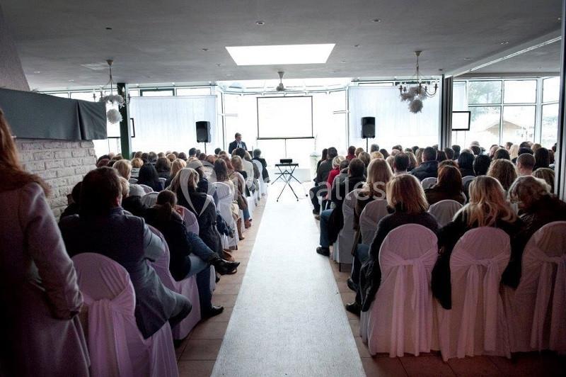 Salle de conférence remplie de participants assis, écoutant un intervenant debout devant un écran de présentation.
