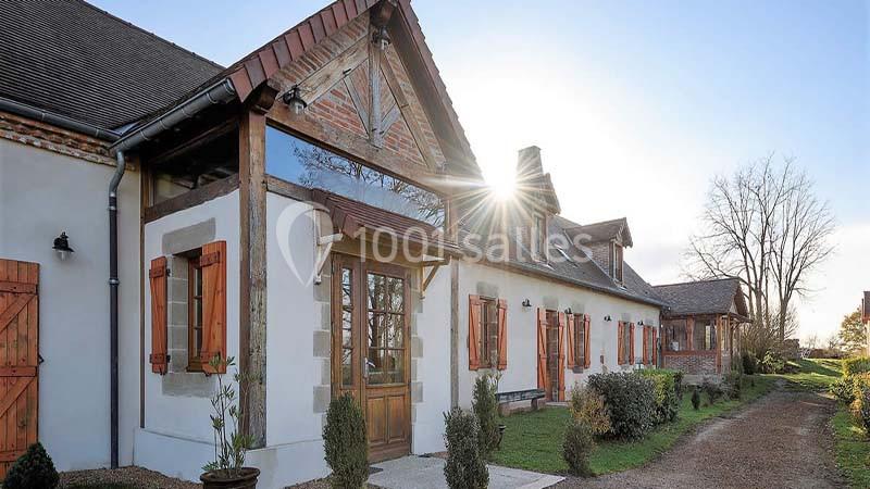 Façade d'une maison en briques et bois avec fenêtres à volets, entourée de végétation et baignée de lumière naturelle.
