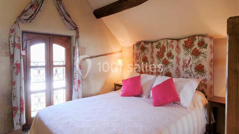 Lit double avec tête de lit fleurie, draps blancs et coussins roses dans une chambre mansardée lumineuse.