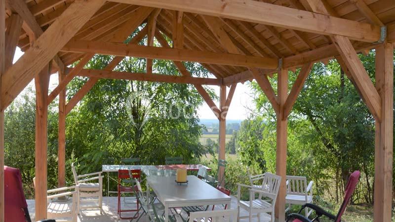 Pergola en bois avec table et chaises, entourée de verdure et offrant une vue dégagée sur la campagne.
