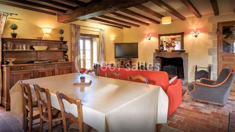 Salle à manger rustique avec table en bois, cheminée, canapé rouge, meubles anciens et poutres apparentes.
