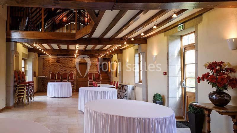 Salle de réception lumineuse avec poutres apparentes, tables rondes drapées de blanc et chaises empilées dans un coin.