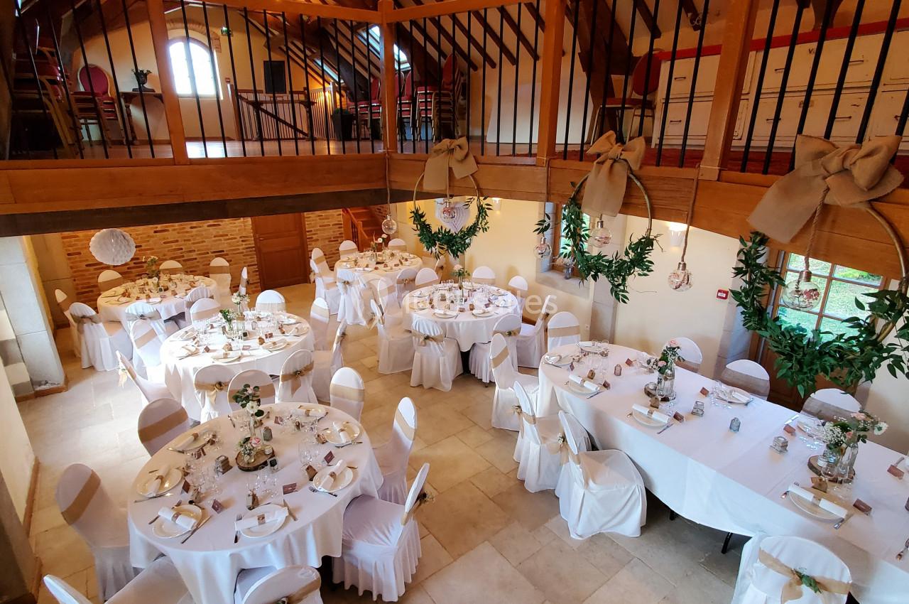 Salle de réception décorée avec des tables rondes dressées, chaises blanches et poutres apparentes en bois.