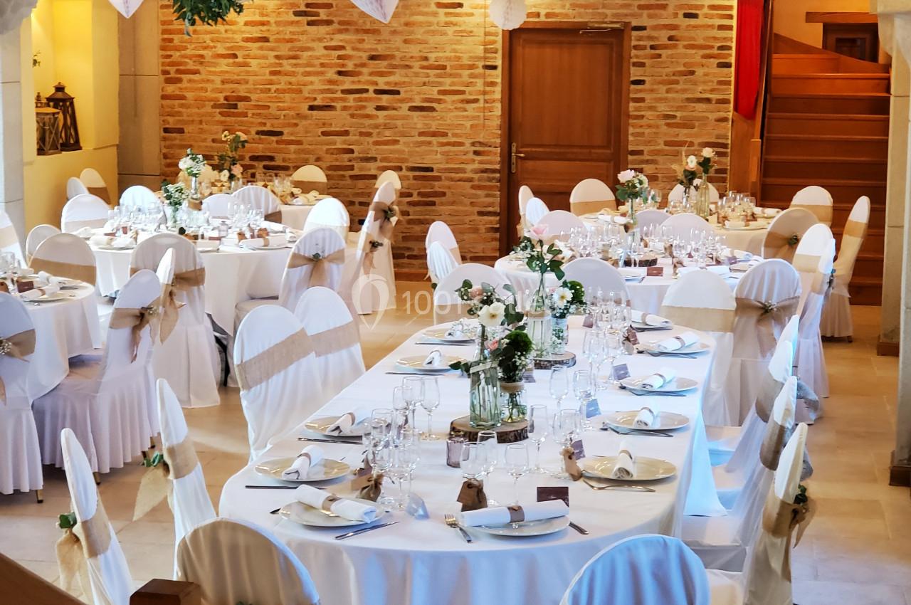 Salle de réception décorée pour un mariage, avec tables rondes dressées, chaises blanches et éclairage chaleureux.