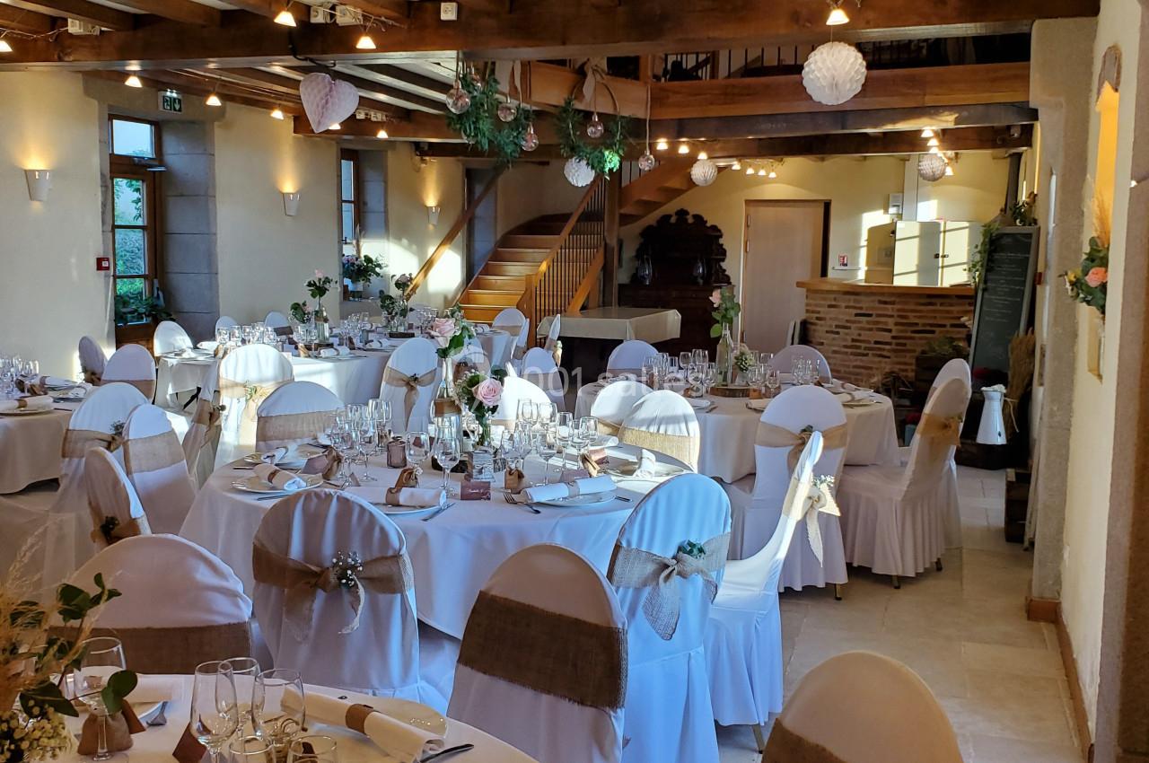 Salle de réception décorée avec des tables rondes dressées, chaises blanches et poutres apparentes au plafond.