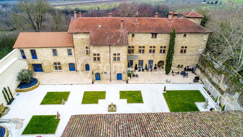Vue aérienne d'un bâtiment en pierre avec une cour aménagée, des pelouses carrées et un paysage rural en arrière-plan.