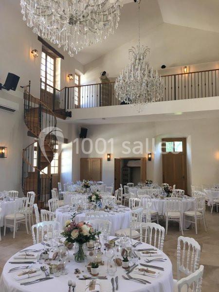 Salle de réception lumineuse avec tables rondes décorées pour un événement, escalier en colimaçon et lustres en cristal.