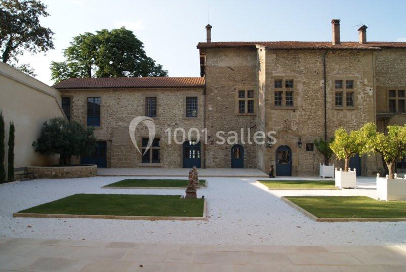 Cour intérieure avec jardin aménagé, pelouse carrée, gravier blanc et bâtiment en pierre avec fenêtres et portes bleues.