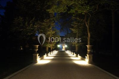 Allée éclairée par des lampes au sol, bordée d'arbres et menant à une entrée illuminée dans l'obscurité.