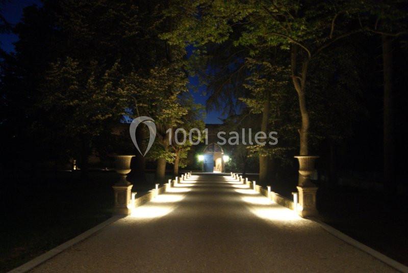 Allée éclairée par des lampes au sol, bordée d'arbres et menant à une entrée illuminée dans l'obscurité.