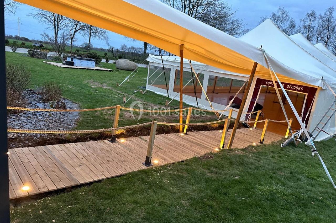 Passerelle en bois éclairée menant à une tente blanche installée sur une pelouse, entourée d'arbres et d'arbustes.