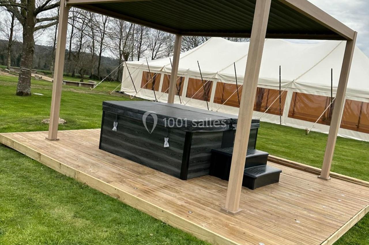Bain à remous noir sous une pergola en bois sur une terrasse en bois, installé dans un jardin avec une tente en arrière-plan.