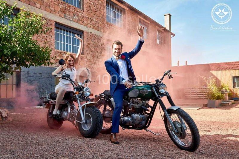 Un homme en costume et une femme en robe blanche sur des motos, entourés de fumée colorée dans une cour ensoleillée.