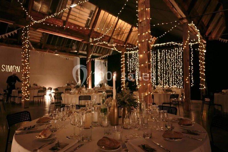 Salle de réception décorée avec des guirlandes lumineuses, des tables rondes dressées et une ambiance chaleureuse.