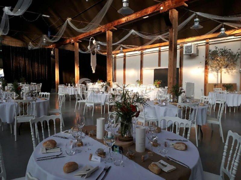 Salle de réception décorée avec des tables rondes dressées, des chaises blanches et des voilages suspendus.
