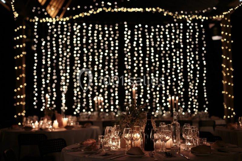 Salle de réception décorée avec des guirlandes lumineuses suspendues et des tables dressées avec bougies et verres.