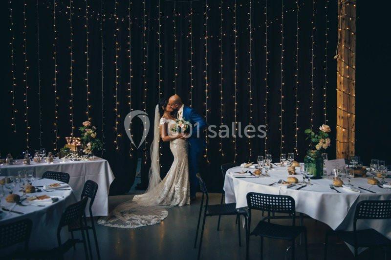 Un couple de mariés s'embrasse dans une salle décorée de guirlandes lumineuses, entourée de tables dressées.