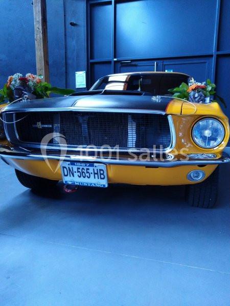 Voiture Ford Mustang jaune et noire exposée en intérieur, ornée de fleurs sur le pare-chocs avant.