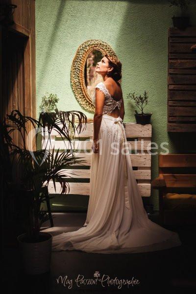 Femme en robe de mariée blanche, debout dans un intérieur lumineux avec des plantes et un miroir en arrière-plan.