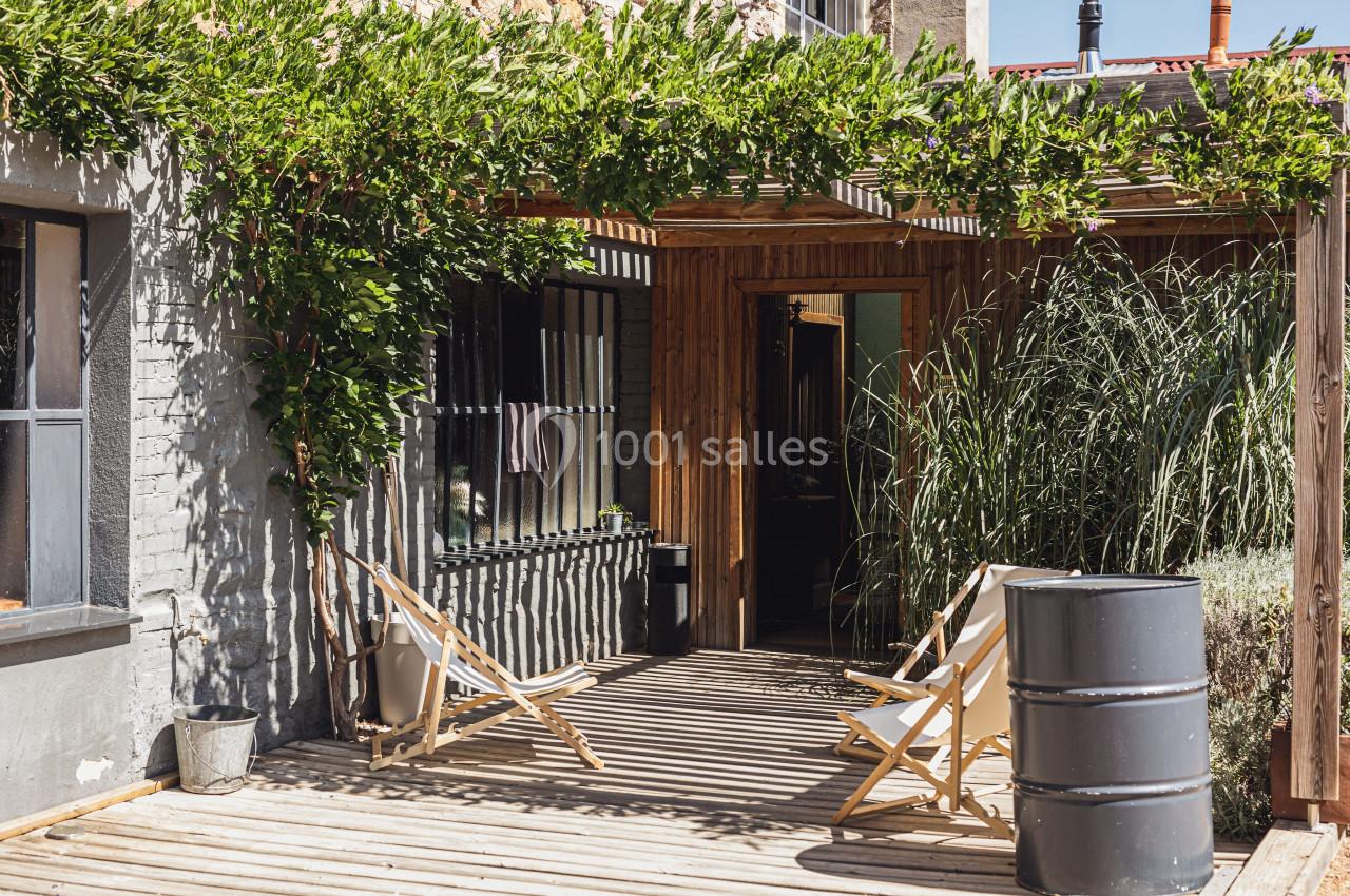 Terrasse en bois avec chaises longues, pergola couverte de verdure et plantes entourant l'entrée d'une maison.