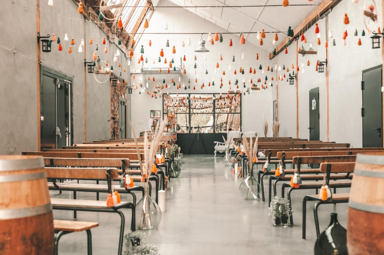 Salle décorée pour un événement, avec bancs en bois, guirlandes suspendues et fleurs dans des vases en verre.