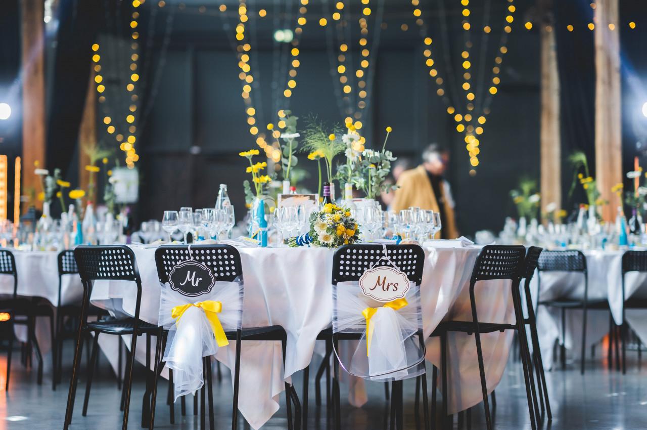Salle décorée pour un mariage avec tables dressées, chaises ornées de panneaux ’Mr’ et ’Mrs’, et guirlandes lumineuses…
