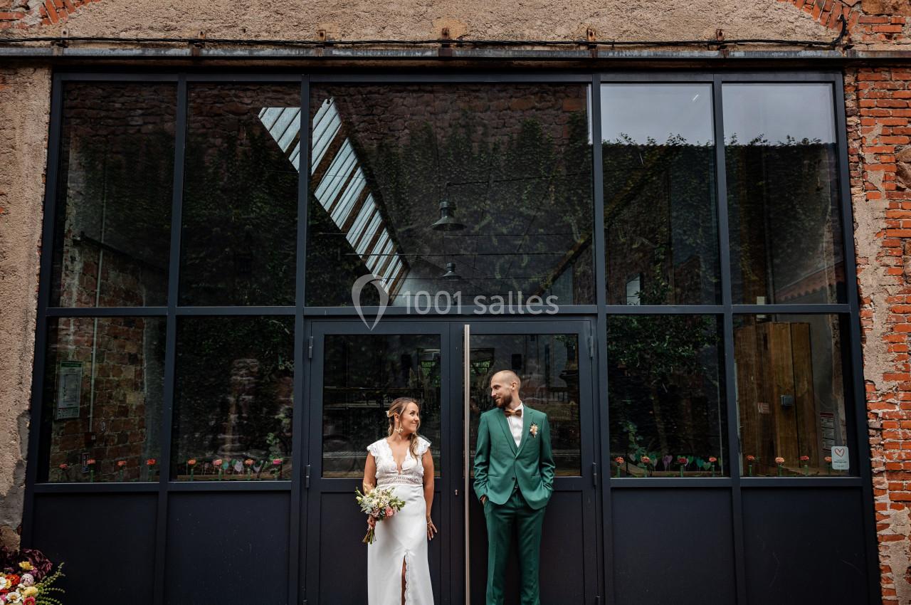 Un couple en tenue de mariage se tient devant une grande porte vitrée dans un bâtiment en briques.
