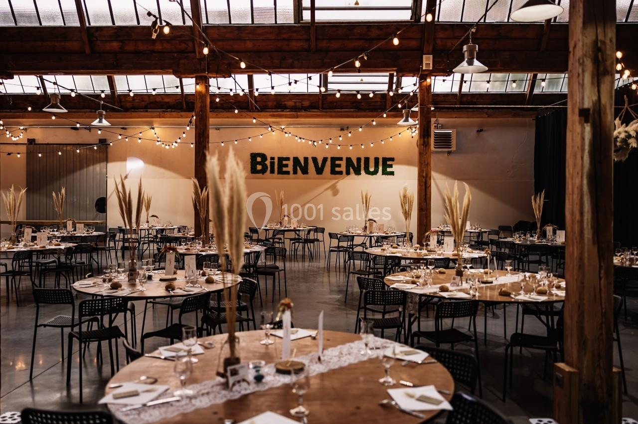 Salle de réception décorée avec des tables rondes, des pampas et des guirlandes lumineuses, avec un mur ’Bienvenue’.