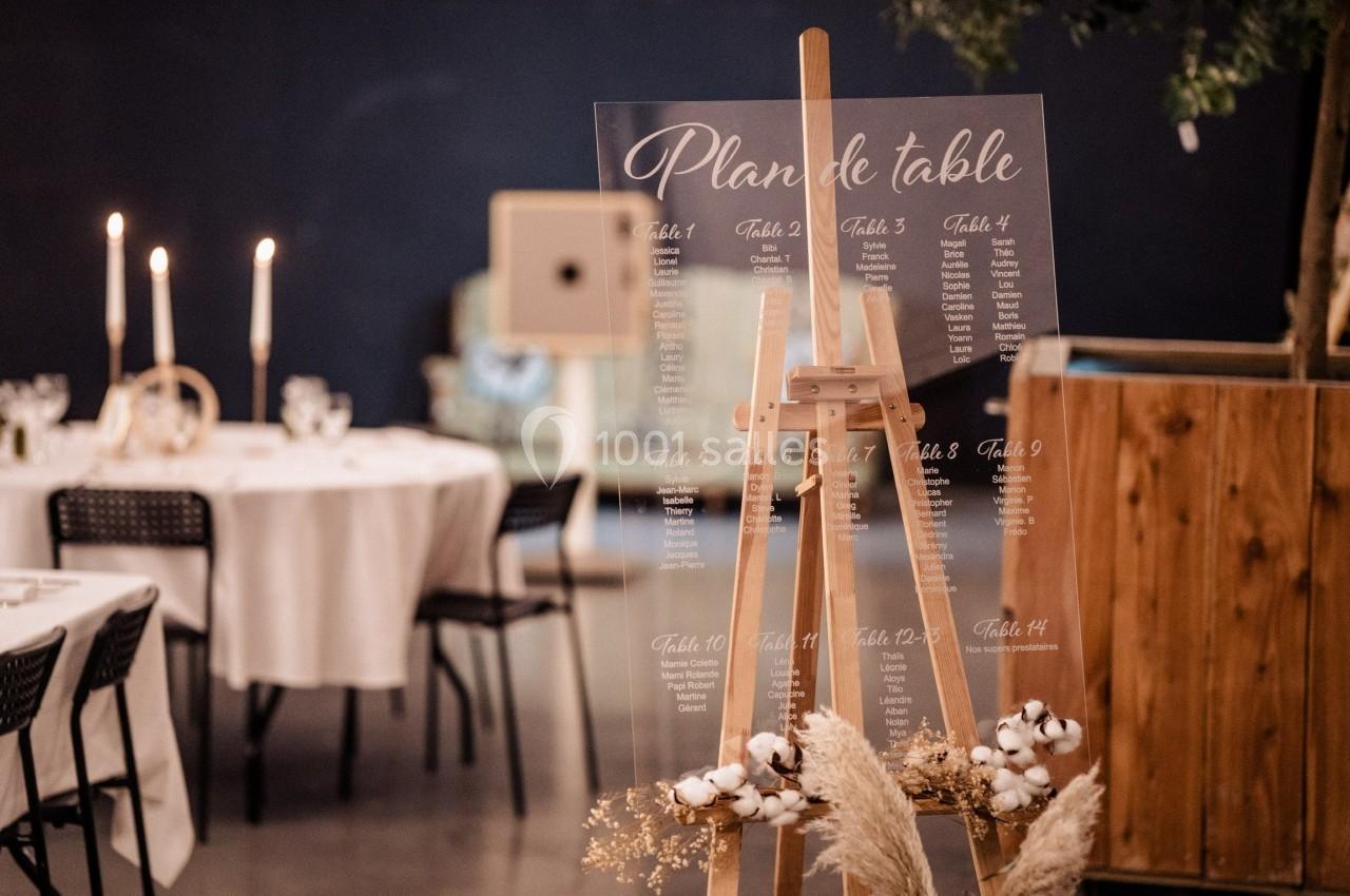 Plan de table transparent sur un chevalet en bois, entouré de décorations florales et de tables dressées.