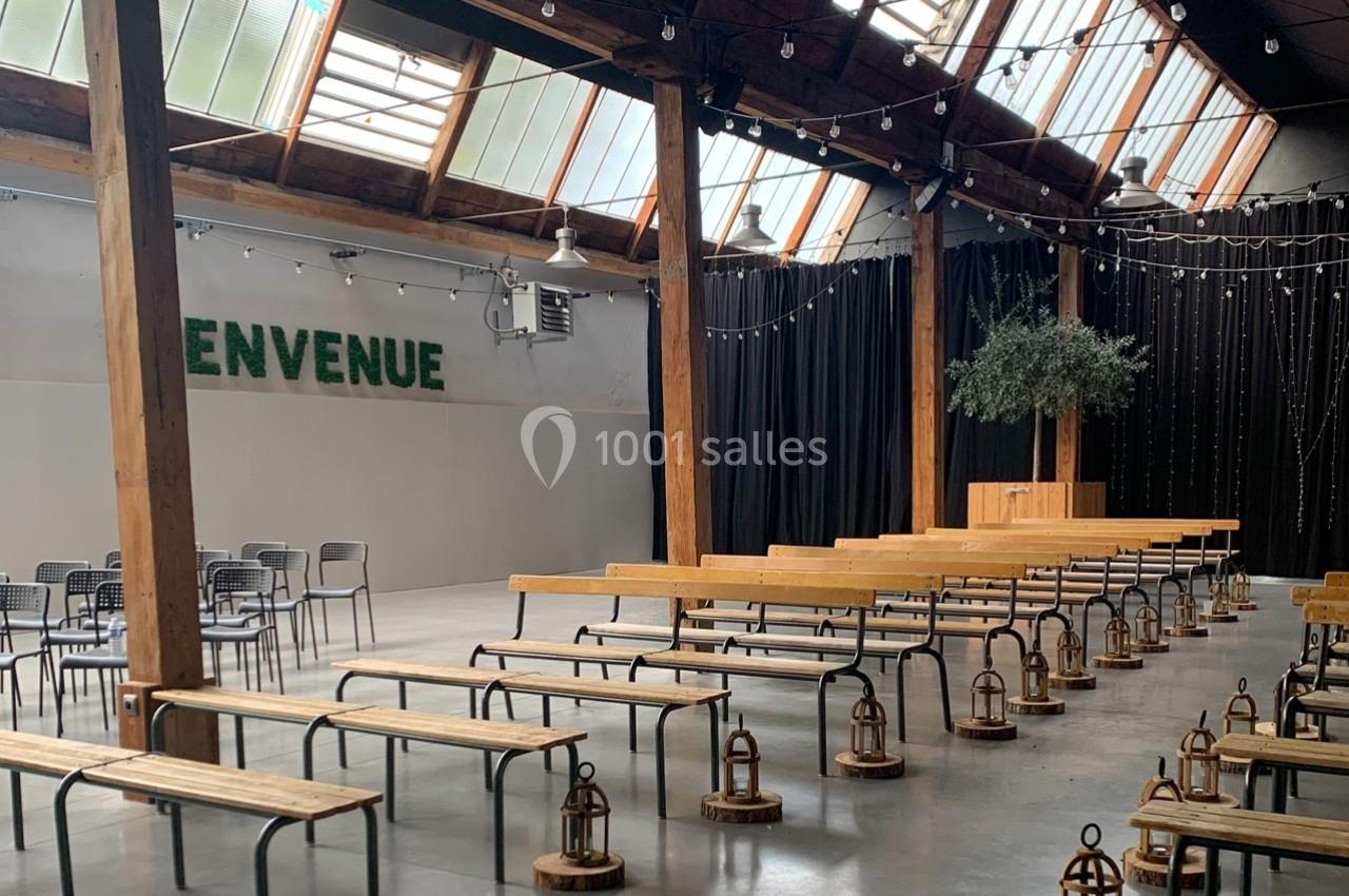 Salle lumineuse avec bancs en bois, guirlandes suspendues, mur végétal ’Bienvenue’ et scène avec un arbre décoratif.