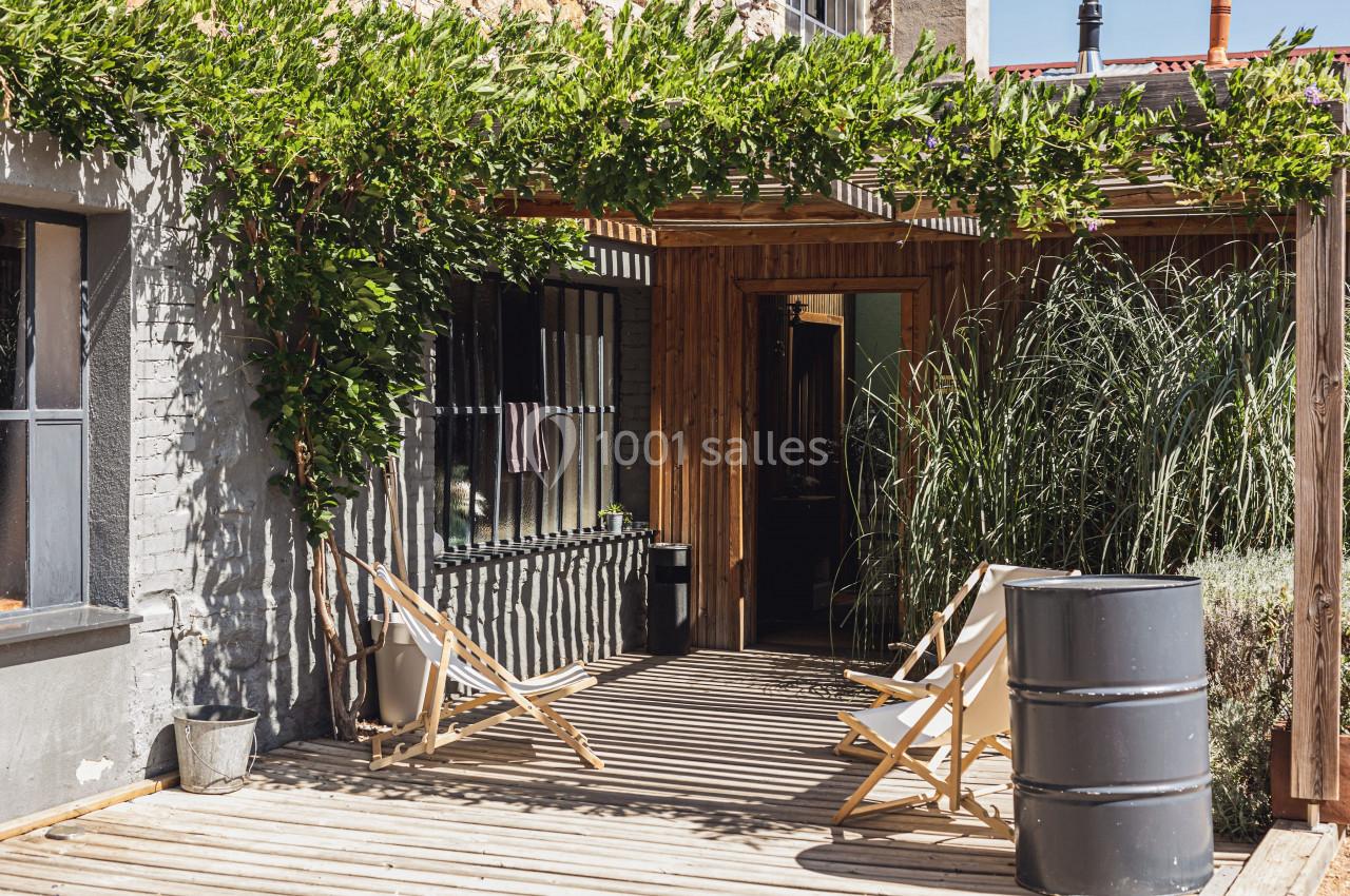 Terrasse en bois avec chaises longues, pergola couverte de végétation et façade en briques avec grandes fenêtres.