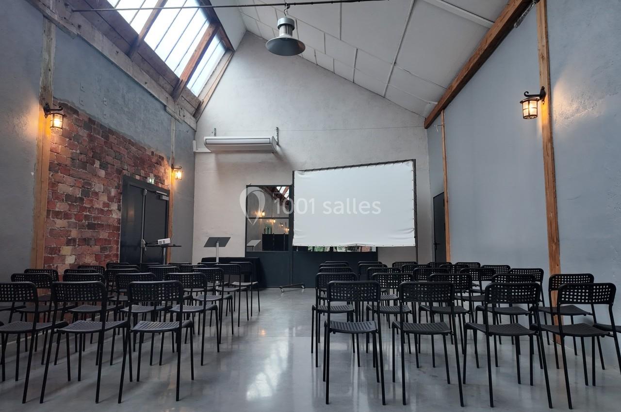 Salle lumineuse avec des rangées de chaises noires, un écran de projection blanc et des murs en briques et béton.
