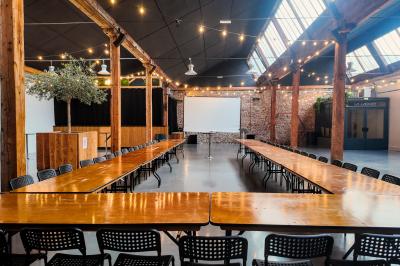 Salle lumineuse avec de grandes tables en bois dressées, chaises noires, décor minimaliste et verrière en hauteur.