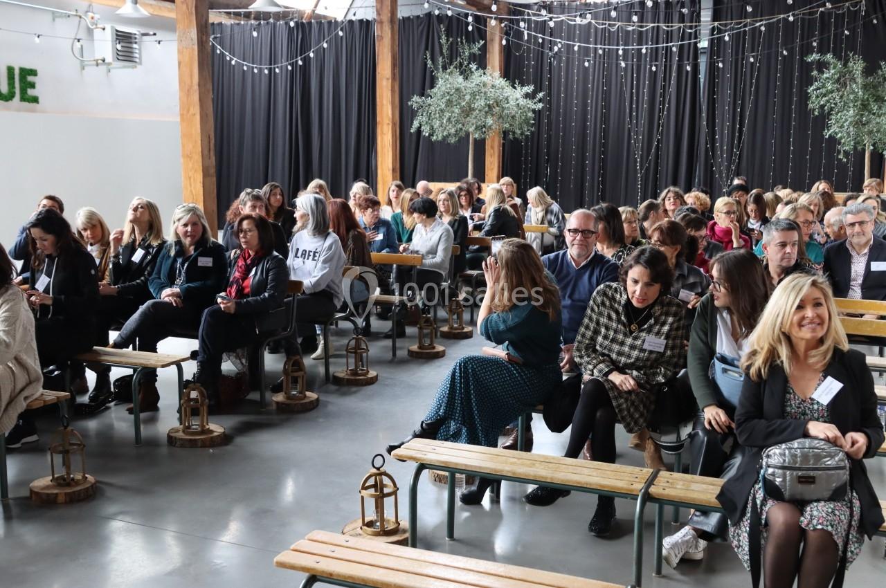 Des personnes assises sur des bancs en bois dans une salle décorée de guirlandes lumineuses et de plantes.