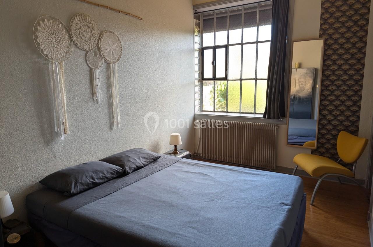 Chambre lumineuse avec un lit double, décoration murale en macramé, grande fenêtre et chaise jaune près d'un miroir.