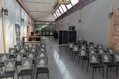 Salle lumineuse avec de grandes tables en bois dressées, chaises noires, décor minimaliste et verrière en hauteur.