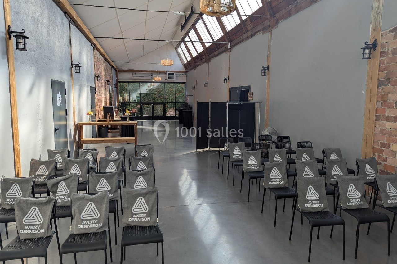 Salle lumineuse avec des rangées de chaises noires alignées, un comptoir en bois et des murs en briques apparentes.