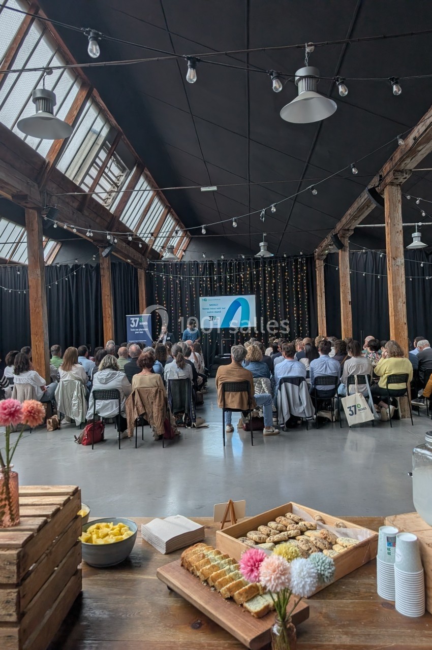 Salle avec un public assis écoutant une présentation, tables au premier plan avec collations et décorations florales.
