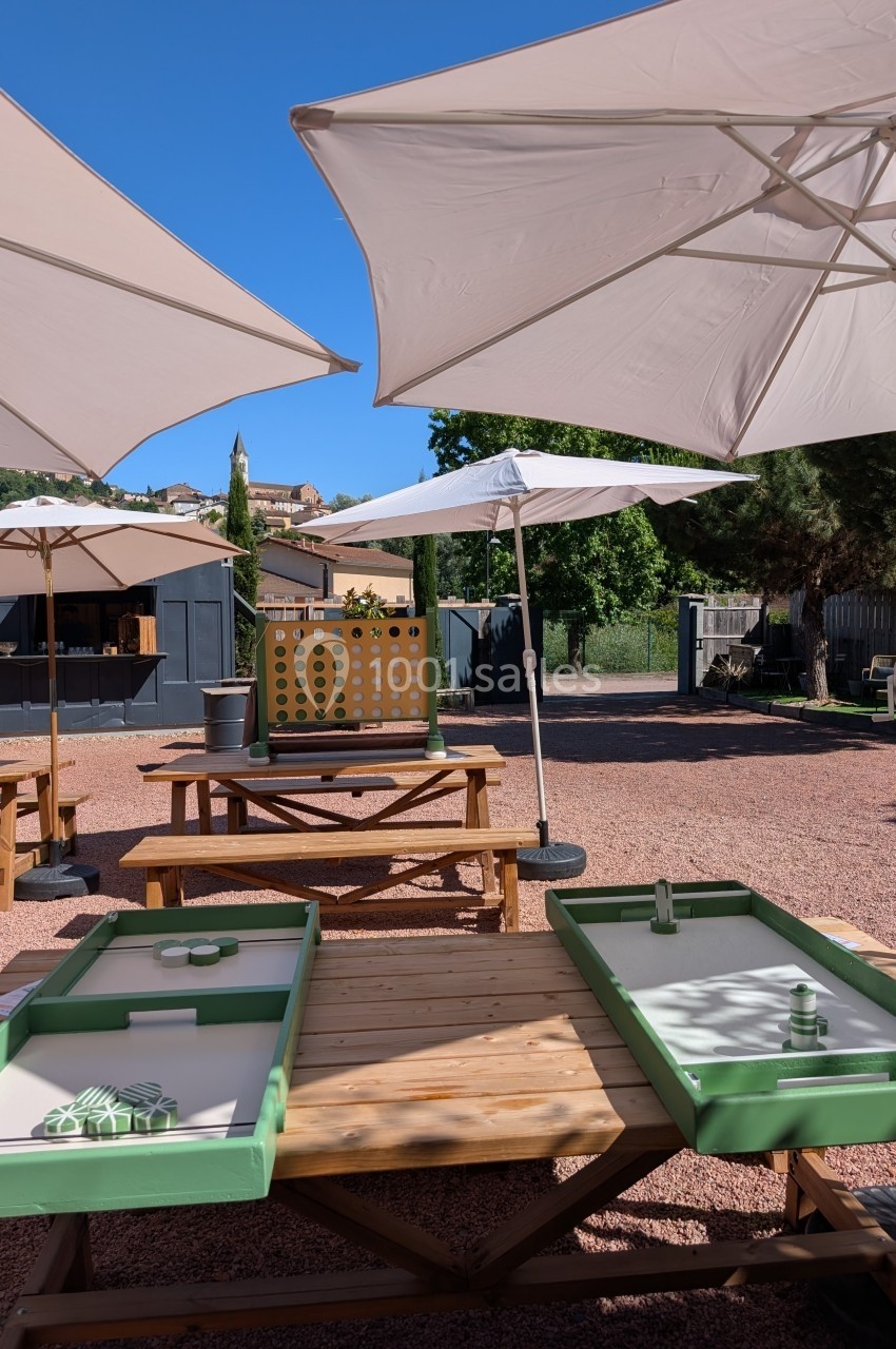 Tables en bois avec jeux de palets, parasols blancs et décor extérieur ensoleillé avec arbres et cabane en arrière-plan.