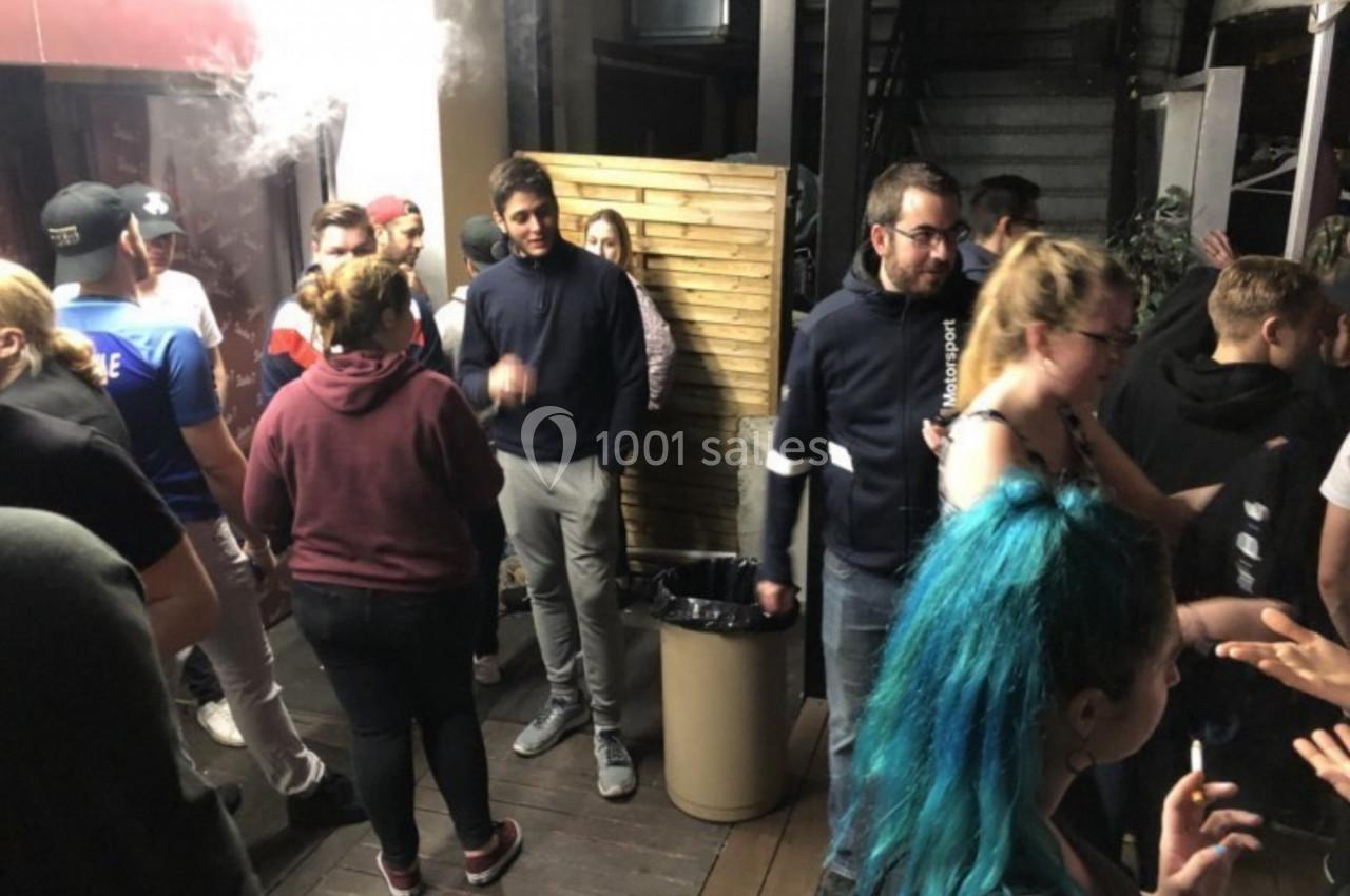 Groupe de personnes discutant et fumant dans un espace extérieur avec un mur en bois et des escaliers en arrière-plan.