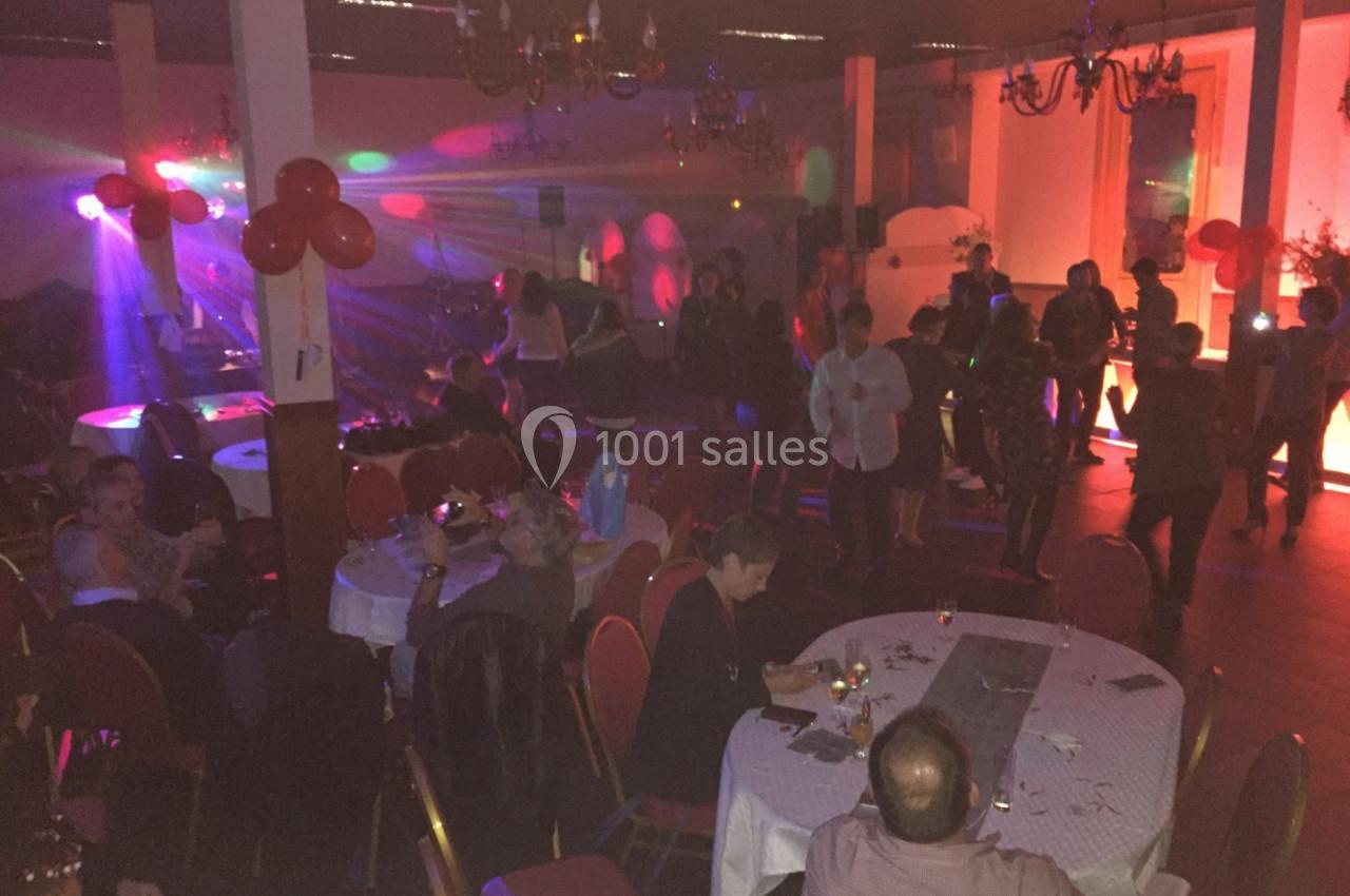 Salle de réception avec des invités assis et debout, éclairée par des lumières colorées et décorée de ballons rouges.