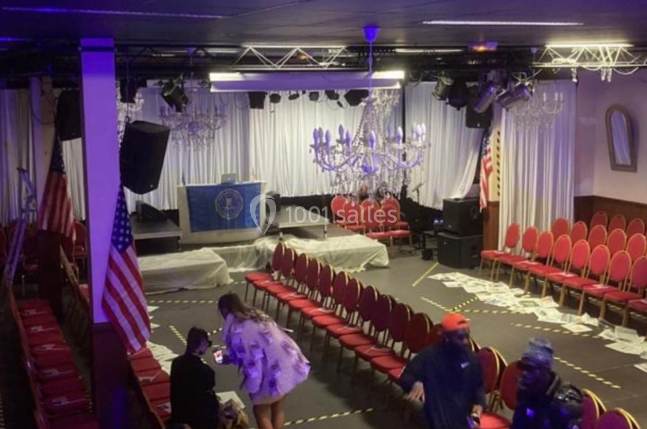 Salle de conférence avec des rangées de chaises rouges, drapeaux américains et un podium sous des lustres.