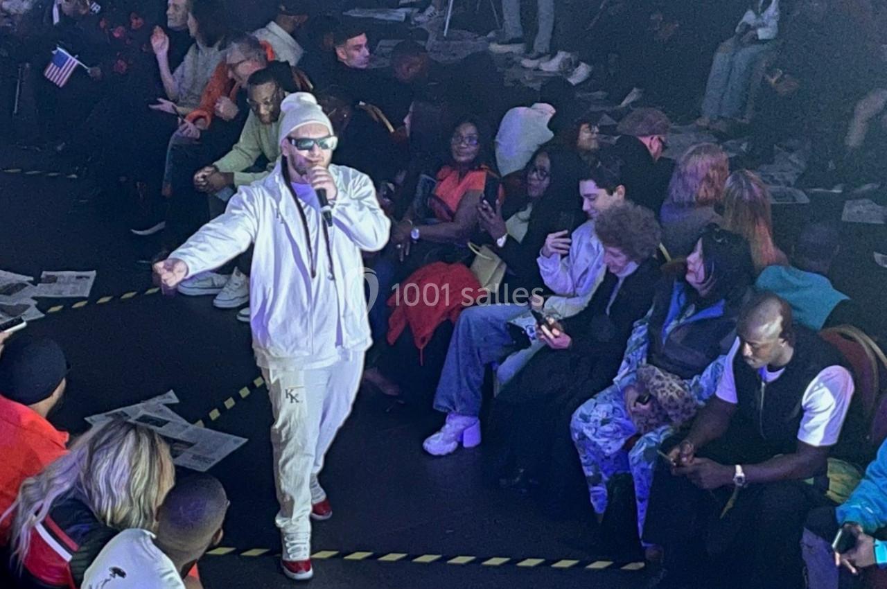 Un artiste en tenue blanche chante au centre d'une salle entourée d'un public assis en cercle.