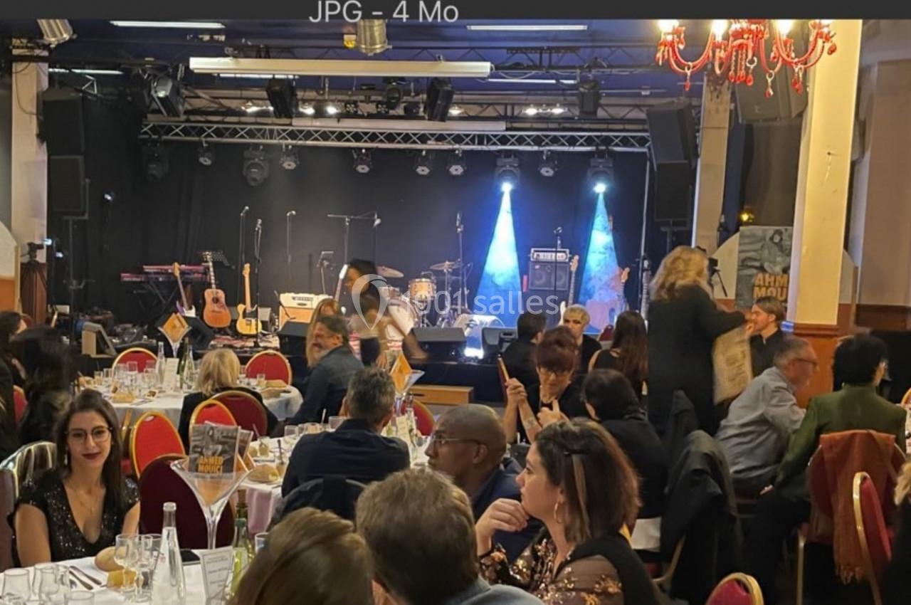Salle de concert avec des tables occupées par des convives, une scène équipée d'instruments et des éclairages en arrière…