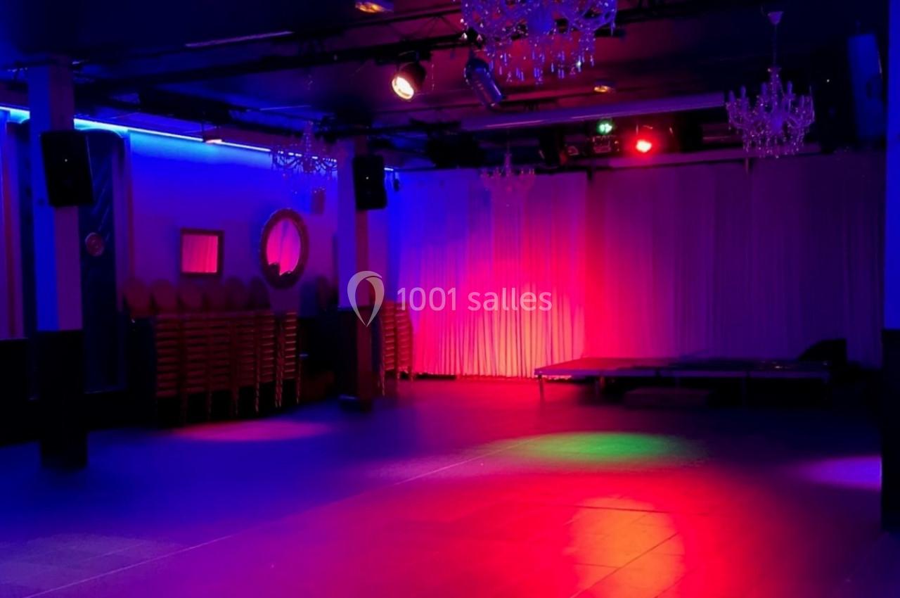 Salle de réception vide éclairée par des lumières colorées, avec des chaises empilées sur le côté et des rideaux blancs.