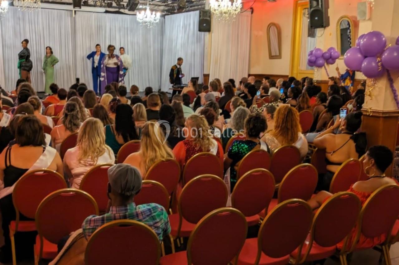 Un public assis dans une salle observe une scène où plusieurs personnes sont debout, avec des ballons violets en décoration.