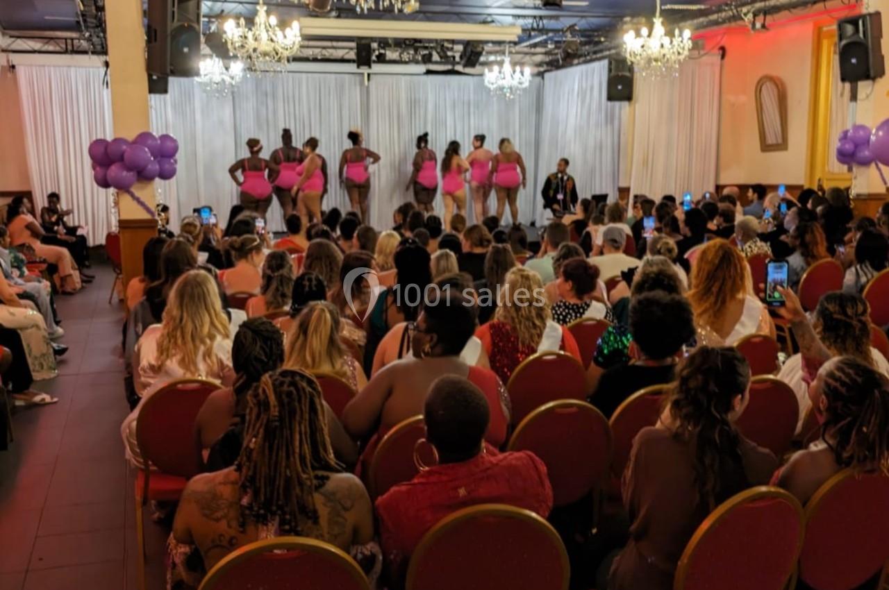 Un groupe de danseurs en tenue rose se produit sur scène devant un public assis dans une salle décorée de ballons violets.