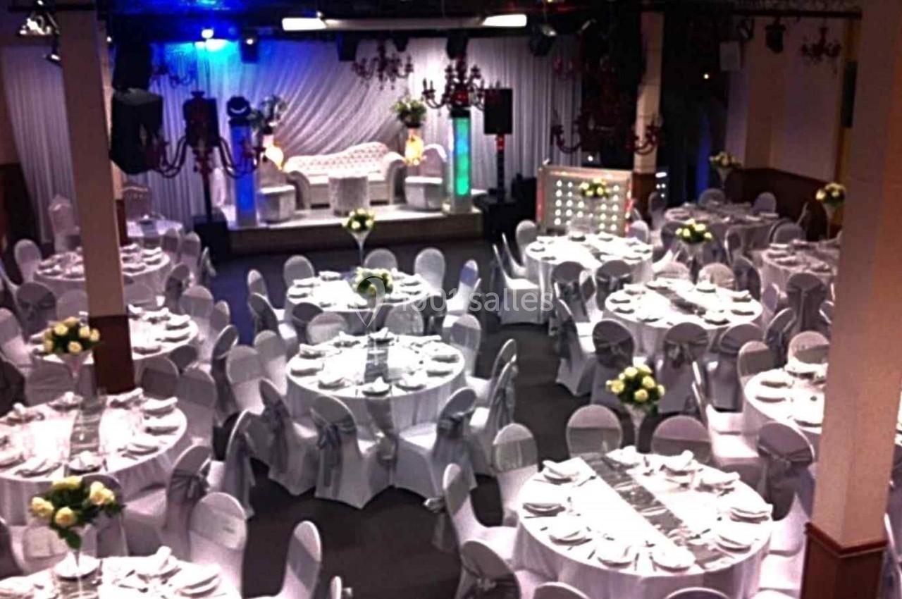 Salle de réception décorée avec des tables rondes dressées, nappes blanches et bouquets de fleurs au centre.